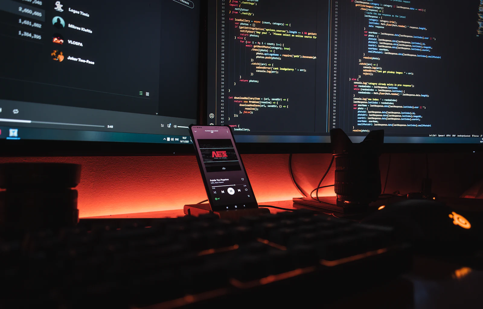 A smartphone on a desk beside monitors displaying lines of code.